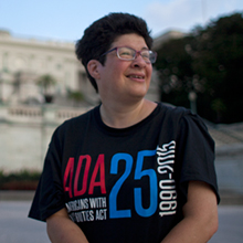 Liz Wientraub stands in front of the United States Capitol. She is wearing glasses and a black t-shirt with 